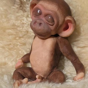 May include: A small, seated monkey figurine with smooth, dark brown skin on its head and limbs, and a lighter, pale beige chest and belly. The monkey has closed eyes, a rounded snout, and prominent, pink-tinted ears, with a textured, realistic appearance.
