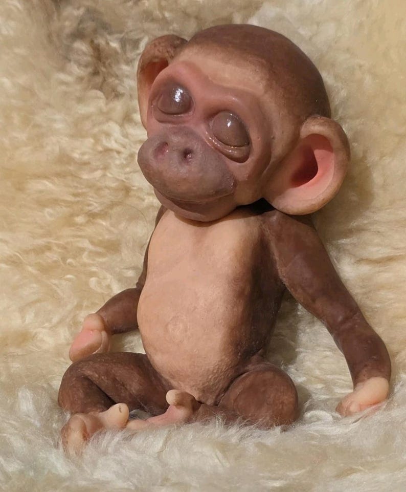 May include: A small, seated monkey figurine with smooth, dark brown skin on its head and limbs, and a lighter, pale beige chest and belly. The monkey has closed eyes, a rounded snout, and prominent, pink-tinted ears, with a textured, realistic appearance.