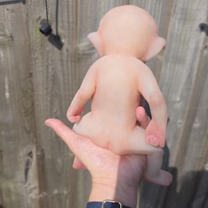 May include: A small, pale, humanoid figurine with pointed ears and a smooth, skin-toned surface. The figure is depicted from the back, showing a rounded head, slender arms, and legs in a seated position. The texture appears matte and uniform.
