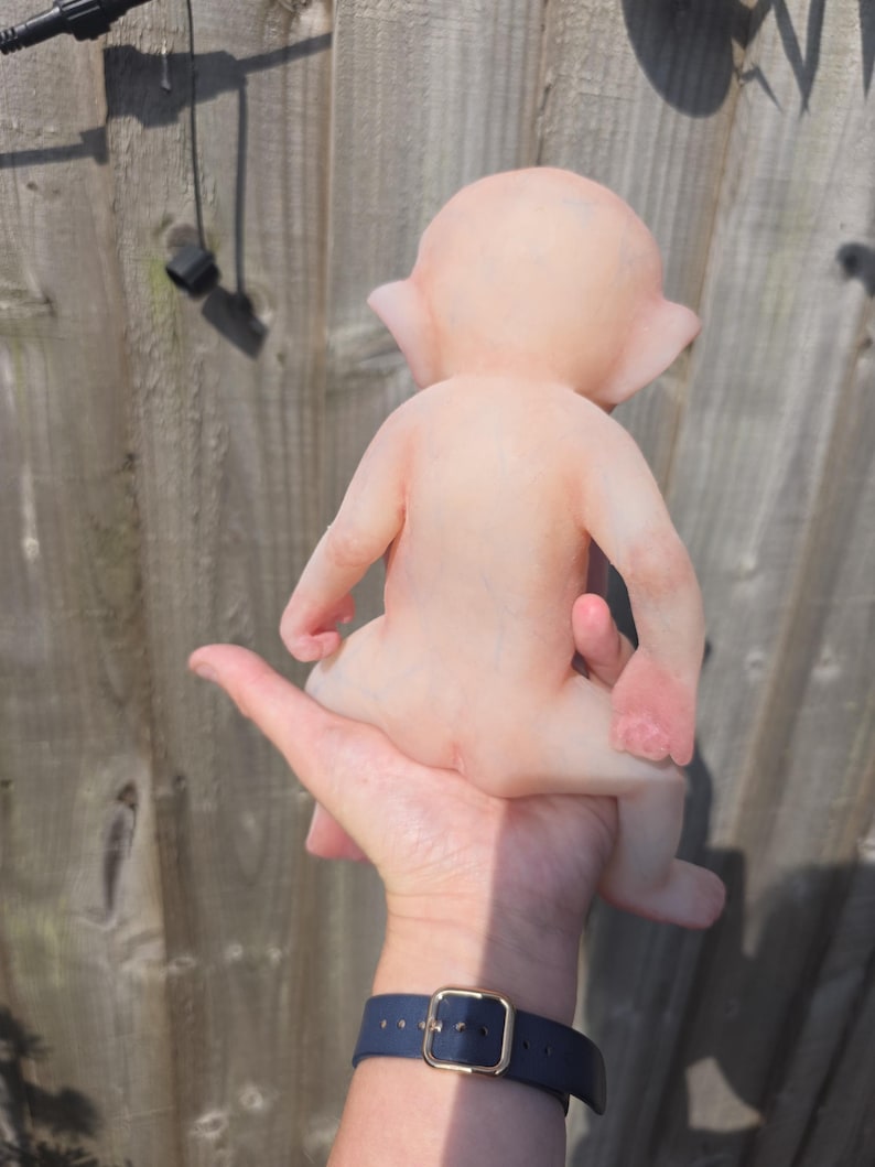 May include: A small, pale, humanoid figurine with pointed ears and a smooth, skin-toned surface. The figure is depicted from the back, showing a rounded head, slender arms, and legs in a seated position. The texture appears matte and uniform.