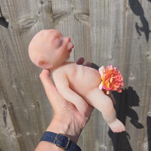 May include: A small, pale, smooth-skinned figurine of a baby with closed eyes and a rounded head. The figure has a light pinkish hue and is positioned with a vibrant, multi-colored orange and pink artificial flower covering the lower torso area.