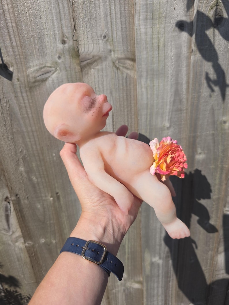 May include: A small, pale, smooth-skinned figurine of a baby with closed eyes and a rounded head. The figure has a light pinkish hue and is positioned with a vibrant, multi-colored orange and pink artificial flower covering the lower torso area.