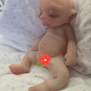 May include: A pale, bald, fantasy-style doll with pointed ears, closed eyes, and visible freckles on the nose and cheeks. The doll has a rounded belly and is positioned in a seated posture with a red flower graphic covering the lower torso area.
