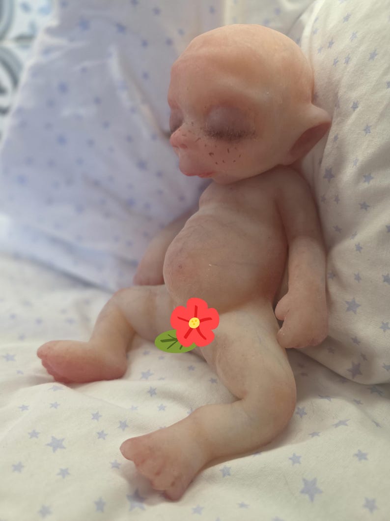 May include: A pale, bald, fantasy-style doll with pointed ears, closed eyes, and visible freckles on the nose and cheeks. The doll has a rounded belly and is positioned in a seated posture with a red flower graphic covering the lower torso area.