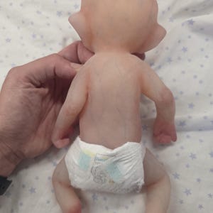 May include: A silicone doll with pale, translucent skin showing subtle blue vein details across the back and limbs. The doll has pointed ears and is wearing a white disposable diaper with light blue and yellow adhesive tabs on the sides.