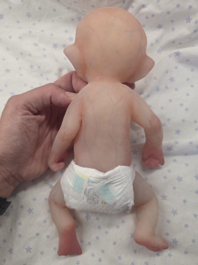 May include: A silicone doll with pale, translucent skin showing subtle blue vein details across the back and limbs. The doll has pointed ears and is wearing a white disposable diaper with light blue and yellow adhesive tabs on the sides.