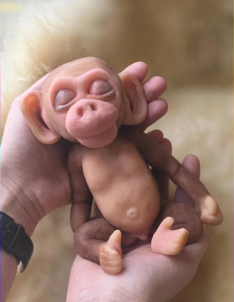 May include: A small, sculpted monkey figurine with a smooth, light brown and tan complexion. The figure features closed eyes, prominent ears, a rounded belly with a visible navel, and articulated limbs, capturing a sleeping, infant-like pose.