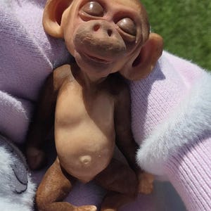 May include: A small, brown, sculpted monkey figurine with closed eyes, prominent ears, and a textured face. The figure has a light-colored belly with a visible navel, dark brown limbs, and light-colored hands and feet, appearing in a resting pose.