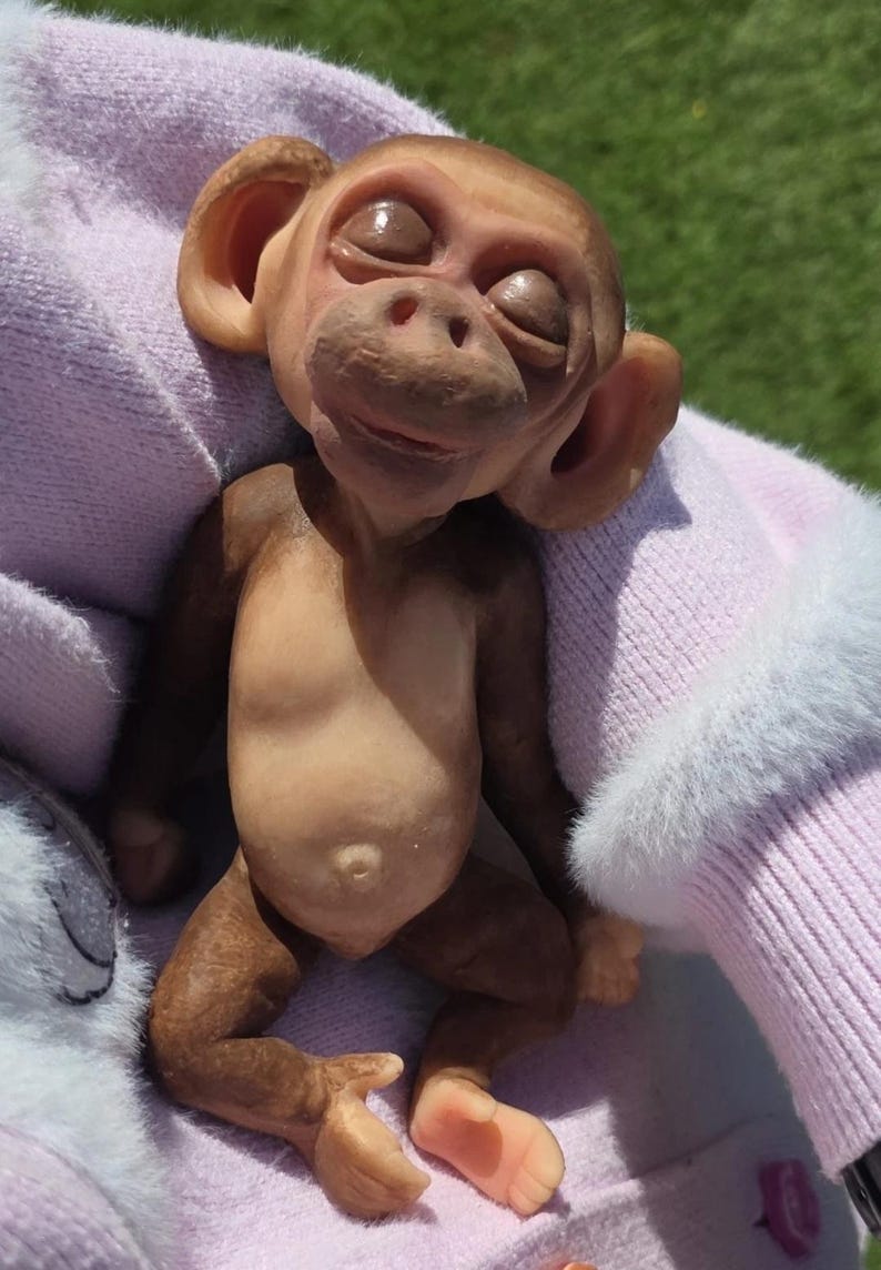 May include: A small, brown, sculpted monkey figurine with closed eyes, prominent ears, and a textured face. The figure has a light-colored belly with a visible navel, dark brown limbs, and light-colored hands and feet, appearing in a resting pose.
