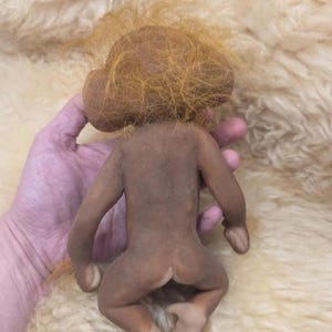 May include: A small, brown, monkey-like figurine with a smooth, matte-textured body and long, wispy, orange-colored hair on its head. The figure has large, rounded ears, slender limbs, and a neutral, unpainted appearance with light-colored feet.