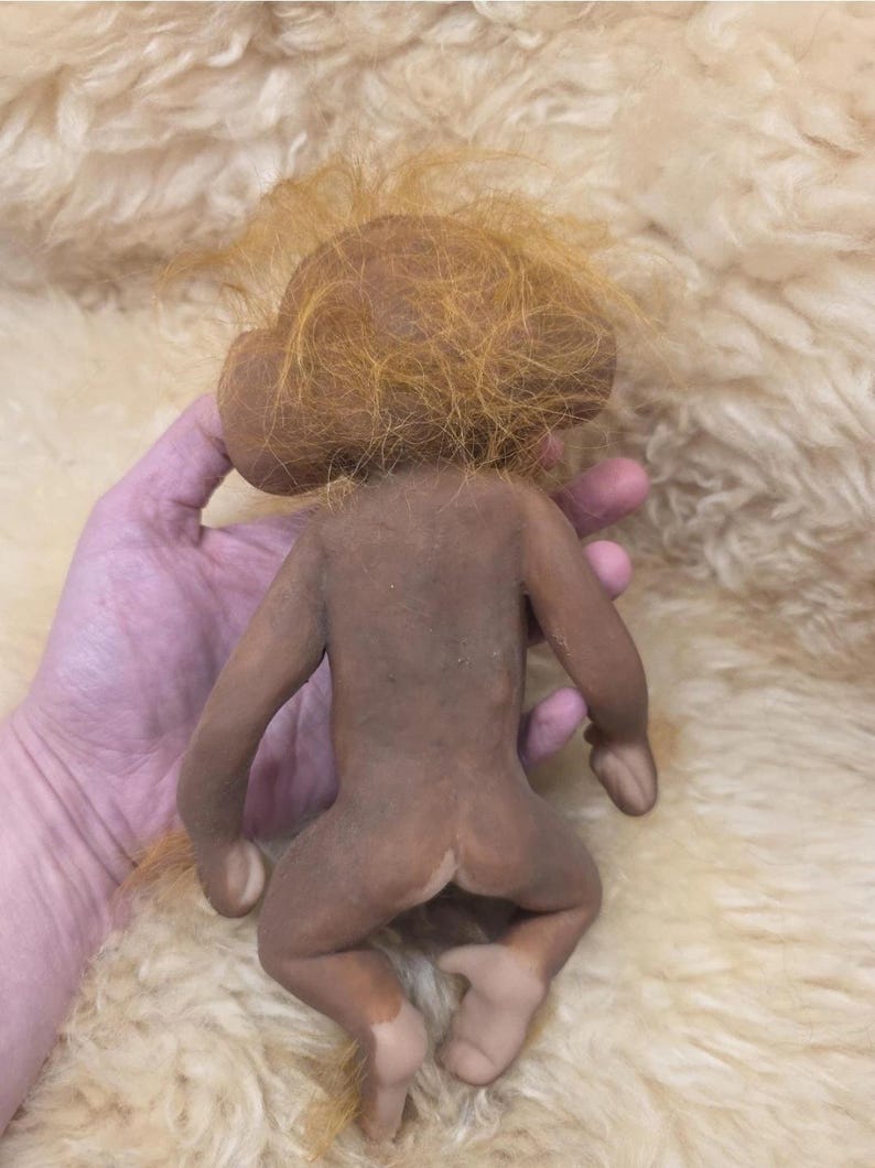 May include: A small, brown, monkey-like figurine with a smooth, matte-textured body and long, wispy, orange-colored hair on its head. The figure has large, rounded ears, slender limbs, and a neutral, unpainted appearance with light-colored feet.