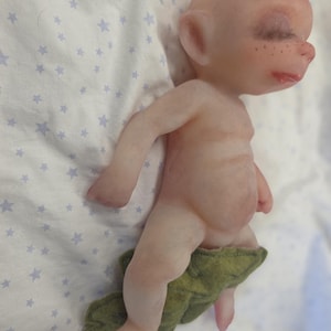 May include: A small, pale, bald figurine with closed eyes, long dark eyelashes, and freckles on the nose. The figure has a rounded belly and is partially covered by a textured, olive green felt leaf-shaped piece around the lower body.