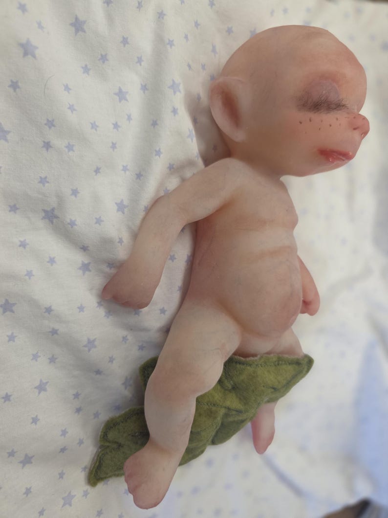 May include: A small, pale, bald figurine with closed eyes, long dark eyelashes, and freckles on the nose. The figure has a rounded belly and is partially covered by a textured, olive green felt leaf-shaped piece around the lower body.