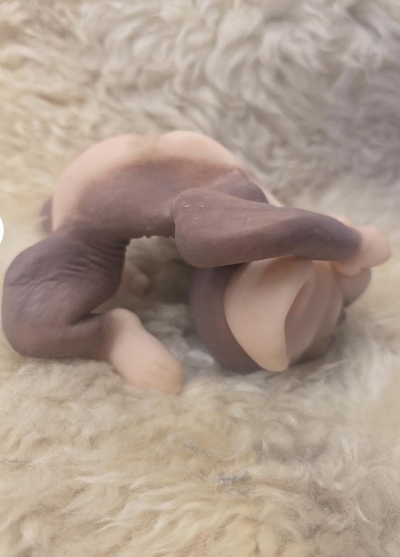 May include: A small, sculpted figurine of a creature in a curled, fetal-like position. The figure features a smooth, matte texture with a two-tone color scheme, combining light beige skin-toned areas with contrasting dark brown patches on the limbs.