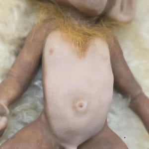 May include: A small monkey figurine with brown skin, a light-colored torso, and a prominent belly button. The figure features textured, ginger-colored fur around the neck and chest area, with detailed facial features including large ears and a wide mouth.