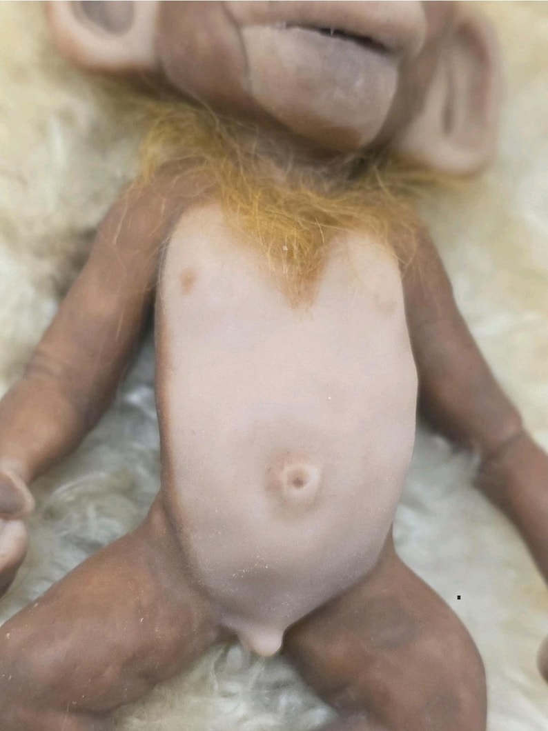 May include: A small monkey figurine with brown skin, a light-colored torso, and a prominent belly button. The figure features textured, ginger-colored fur around the neck and chest area, with detailed facial features including large ears and a wide mouth.