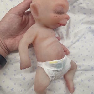 May include: A small, pale-skinned silicone doll with pointed ears, closed eyes, and freckles on the nose. The doll has a smooth, hairless head and is wearing a white disposable diaper with yellow and blue accents around the waist area.