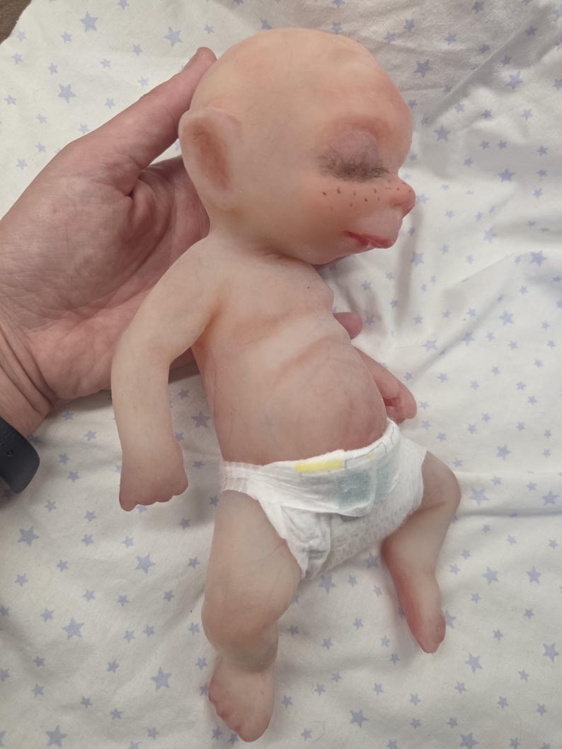 May include: A small, pale-skinned silicone doll with pointed ears, closed eyes, and freckles on the nose. The doll has a smooth, hairless head and is wearing a white disposable diaper with yellow and blue accents around the waist area.