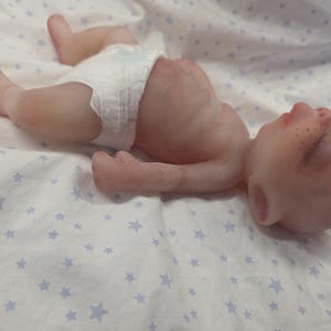 May include: A realistic newborn baby doll with pale skin, closed eyes, and light freckles on the nose. The doll is lying on its back and is wearing a white disposable diaper. The doll has detailed skin texture, small ears, and a rounded belly.