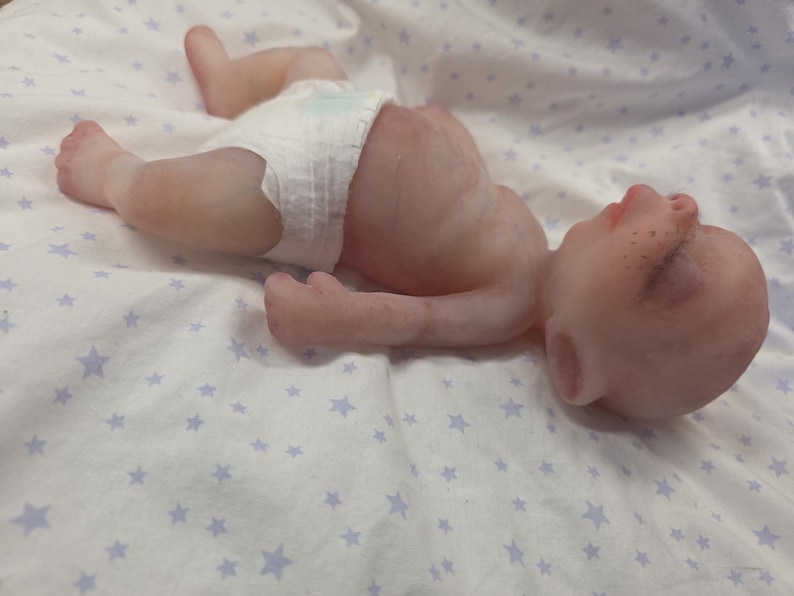 May include: A realistic newborn baby doll with pale skin, closed eyes, and light freckles on the nose. The doll is lying on its back and is wearing a white disposable diaper. The doll has detailed skin texture, small ears, and a rounded belly.