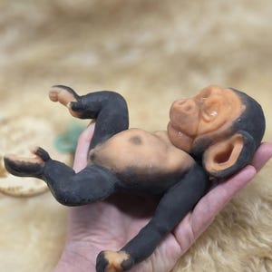 May include: A small, sculpted figurine of a baby chimpanzee with a light peach-colored face, chest, and feet, contrasted by dark black limbs, ears, and head. The figure is depicted in a reclining position with its arms and legs slightly bent.