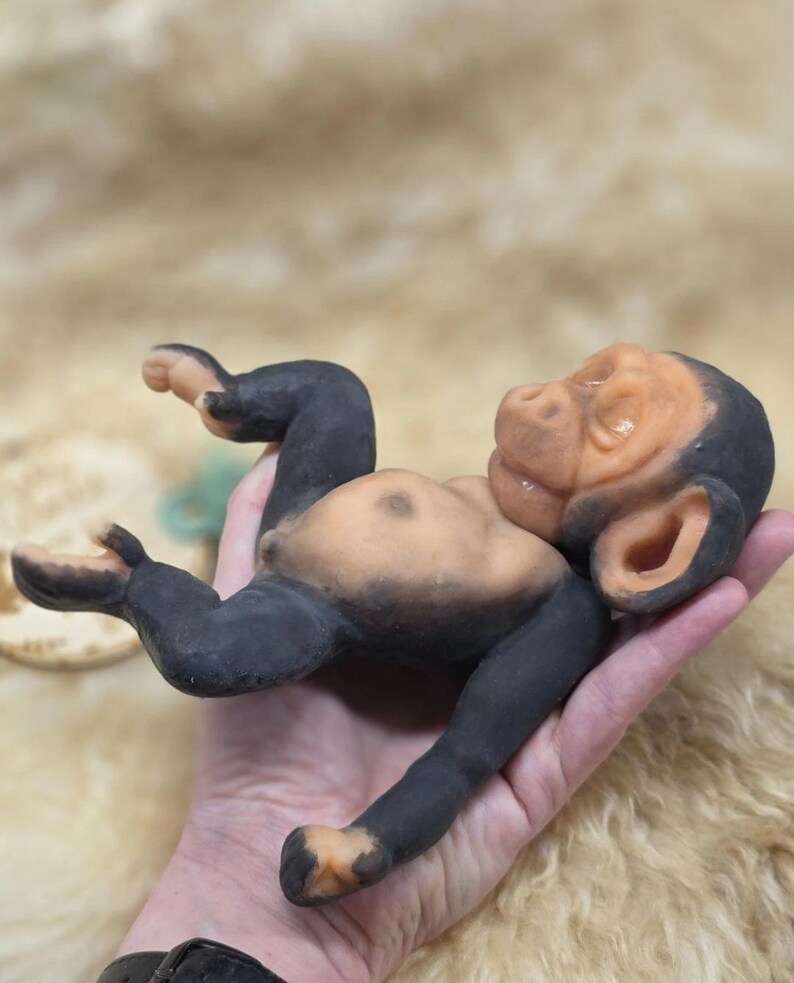 May include: A small, sculpted figurine of a baby chimpanzee with a light peach-colored face, chest, and feet, contrasted by dark black limbs, ears, and head. The figure is depicted in a reclining position with its arms and legs slightly bent.