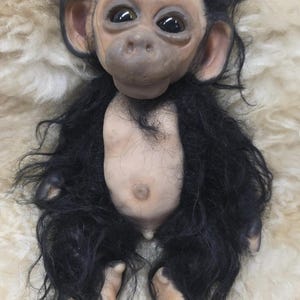 May include: A realistic chimpanzee doll with a light-toned face, ears, hands, and feet. The doll features large, dark, glossy eyes, a prominent nose, and a small mouth. It has a light-toned torso and is covered in long, shaggy black synthetic fur.
