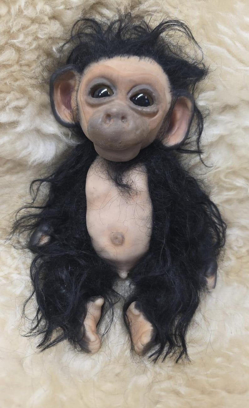 May include: A realistic chimpanzee doll with a light-toned face, ears, hands, and feet. The doll features large, dark, glossy eyes, a prominent nose, and a small mouth. It has a light-toned torso and is covered in long, shaggy black synthetic fur.