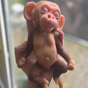 May include: A small, sculpted monkey figurine with a smooth, matte finish. The figure features a light tan face, ears, and torso, transitioning into darker brown limbs. The monkey has closed eyes, a rounded nose, and a visible navel on its belly.