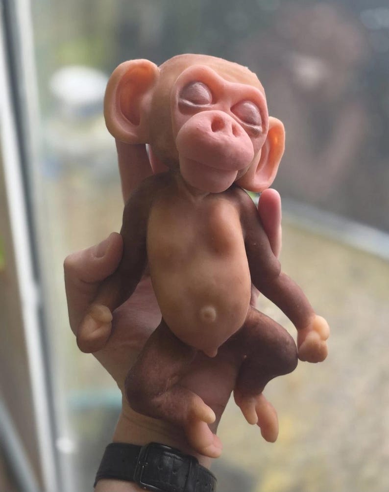 May include: A small, sculpted monkey figurine with a smooth, matte finish. The figure features a light tan face, ears, and torso, transitioning into darker brown limbs. The monkey has closed eyes, a rounded nose, and a visible navel on its belly.
