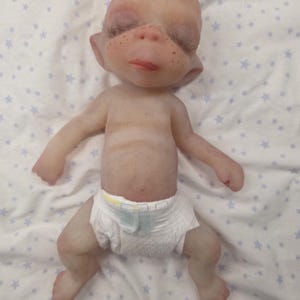 May include: A small, pale-skinned fantasy creature doll with a bald head, pointed ears, and freckles across the nose. The figure has closed eyes with long dark eyelashes, a small mouth, and is wearing a white disposable diaper with yellow accents.