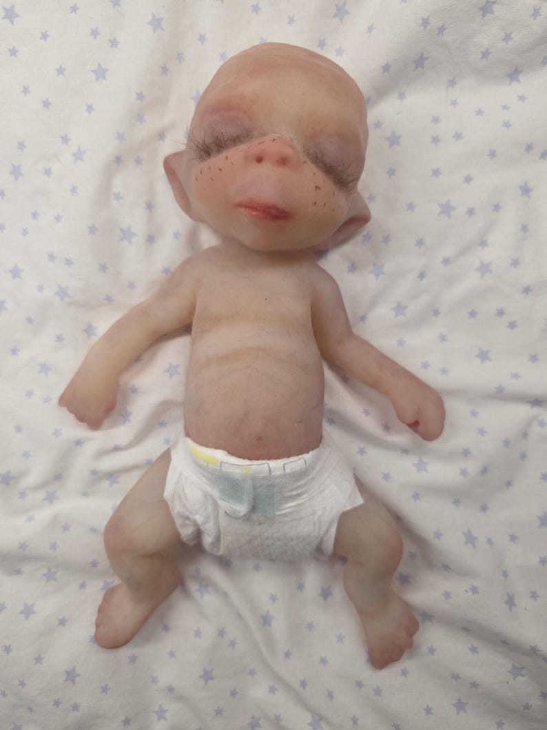 May include: A small, pale-skinned fantasy creature doll with a bald head, pointed ears, and freckles across the nose. The figure has closed eyes with long dark eyelashes, a small mouth, and is wearing a white disposable diaper with yellow accents.