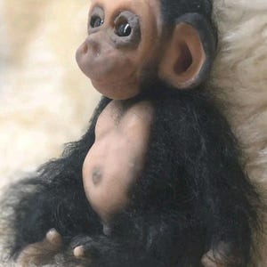 May include: A small, realistic figurine of a baby chimpanzee with a light-toned, smooth face, ears, and belly. The figure is covered in long, shaggy black fur on its head, arms, and back, with dark, expressive eyes and a rounded, protruding snout.