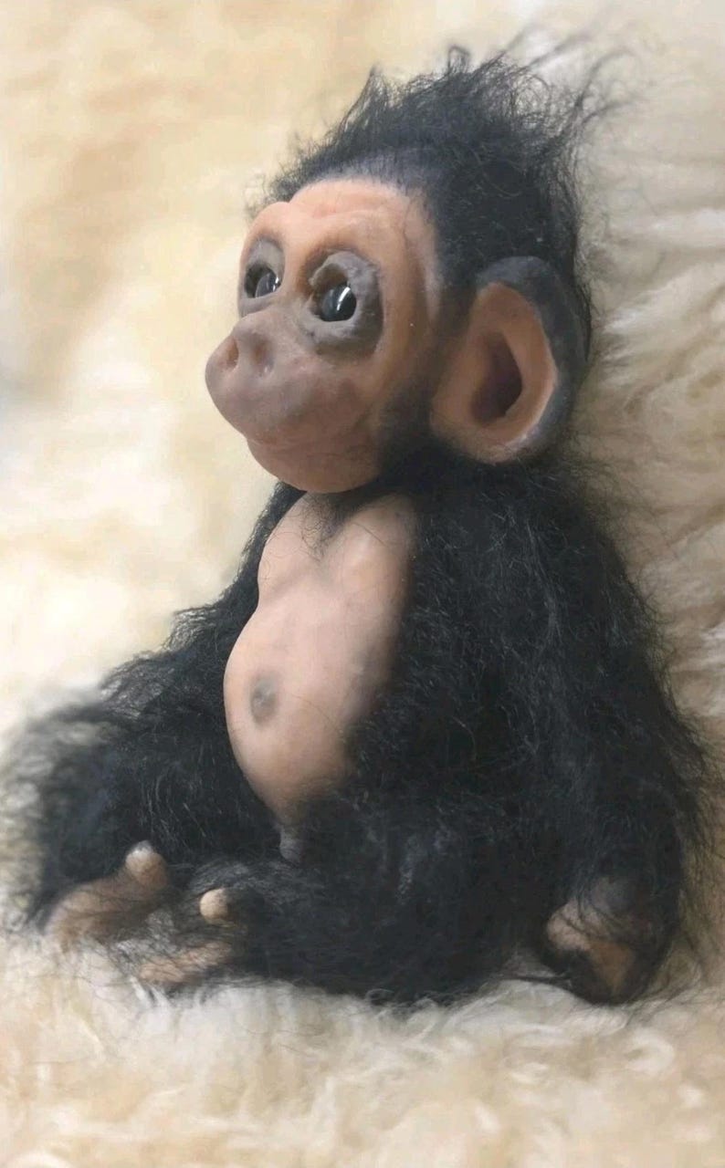 May include: A small, realistic figurine of a baby chimpanzee with a light-toned, smooth face, ears, and belly. The figure is covered in long, shaggy black fur on its head, arms, and back, with dark, expressive eyes and a rounded, protruding snout.
