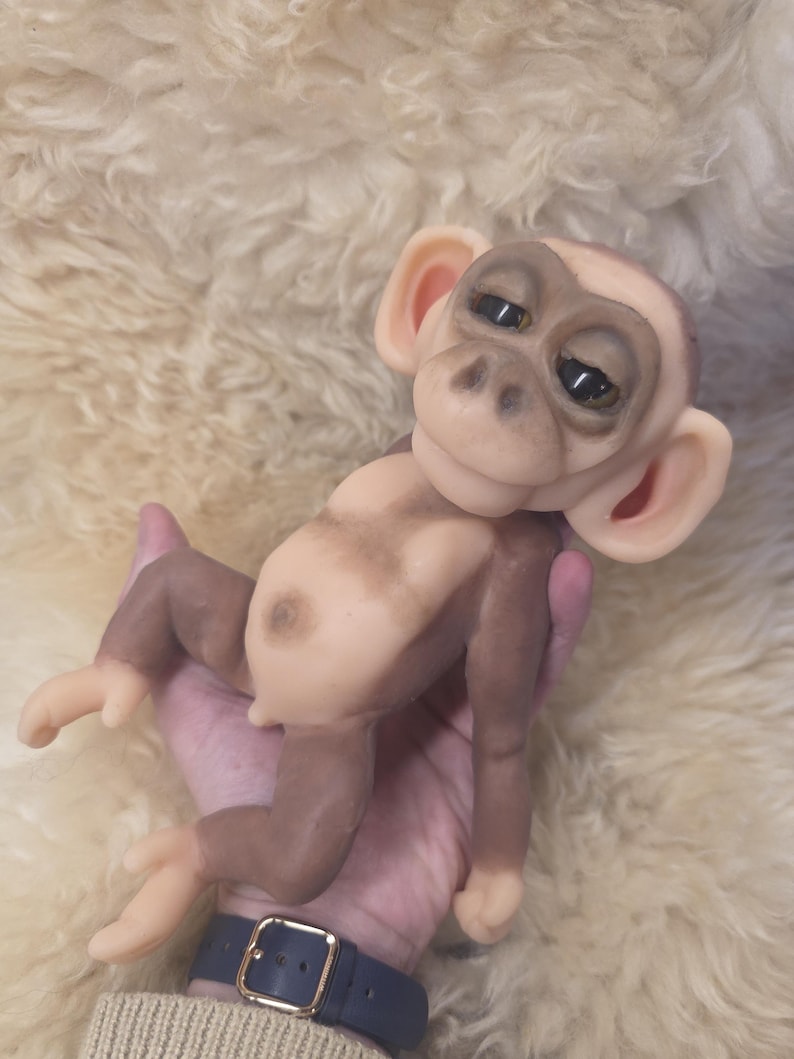 May include: A small, sculpted monkey figurine with a light beige face, ears, and belly, contrasted by dark brown limbs and back. The figure features large, dark, expressive eyes, a rounded nose, and a smooth, matte texture throughout its body.