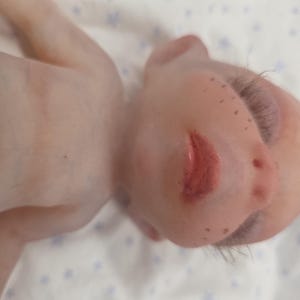 May include: A realistic baby doll with pale skin, light brown freckles across the nose and cheeks, and painted red lips. The doll features long, dark eyelashes, visible veins on the chest, and fine hair, presented in a close-up view.