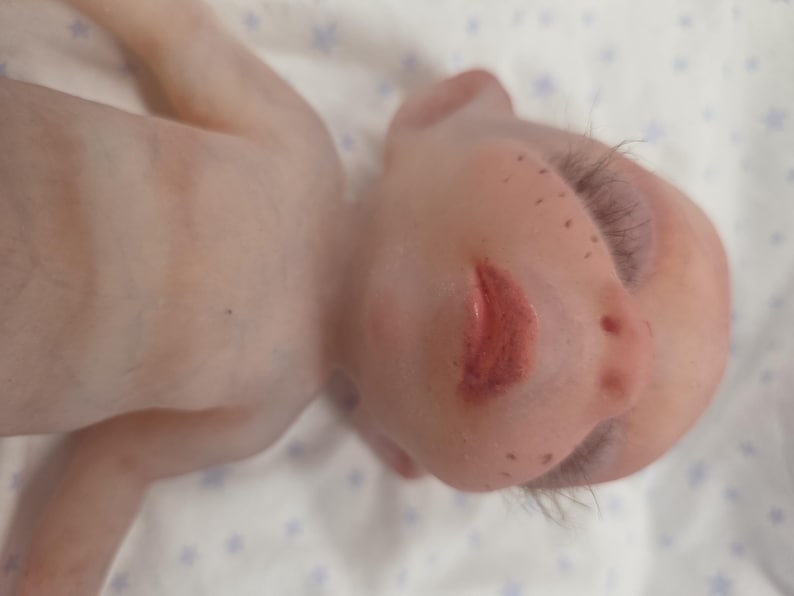 May include: A realistic baby doll with pale skin, light brown freckles across the nose and cheeks, and painted red lips. The doll features long, dark eyelashes, visible veins on the chest, and fine hair, presented in a close-up view.