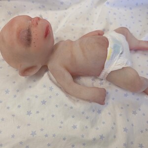 May include: A realistic silicone baby doll with pale skin, closed eyes, dark eyelashes, and freckles across the nose. The doll has pointed ears, a small mouth, and a thin torso. It is wearing a white disposable diaper with a yellow and blue pattern.