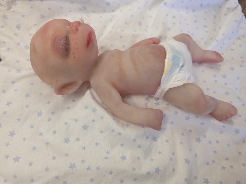 May include: A realistic silicone baby doll with pale skin, closed eyes, dark eyelashes, and freckles across the nose. The doll has pointed ears, a small mouth, and a thin torso. It is wearing a white disposable diaper with a yellow and blue pattern.