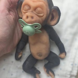 May include: A small, realistic monkey figurine with dark gray limbs and a light tan torso, face, and ears. The monkey has closed eyes and a light green pacifier in its mouth. The figure is sculpted with detailed facial features and a small belly button.