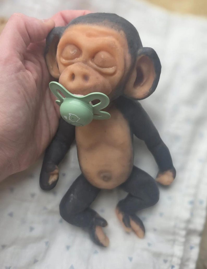 May include: A small, realistic monkey figurine with dark gray limbs and a light tan torso, face, and ears. The monkey has closed eyes and a light green pacifier in its mouth. The figure is sculpted with detailed facial features and a small belly button.