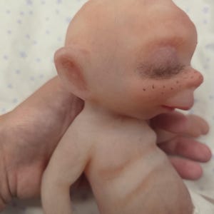 May include: A small, bald, pale-skinned doll with prominent ears, closed eyes with dark eyelashes, and visible freckles across the nose bridge. The doll has a rounded head, a small mouth, and a torso with subtle brown shading, wearing a white diaper.
