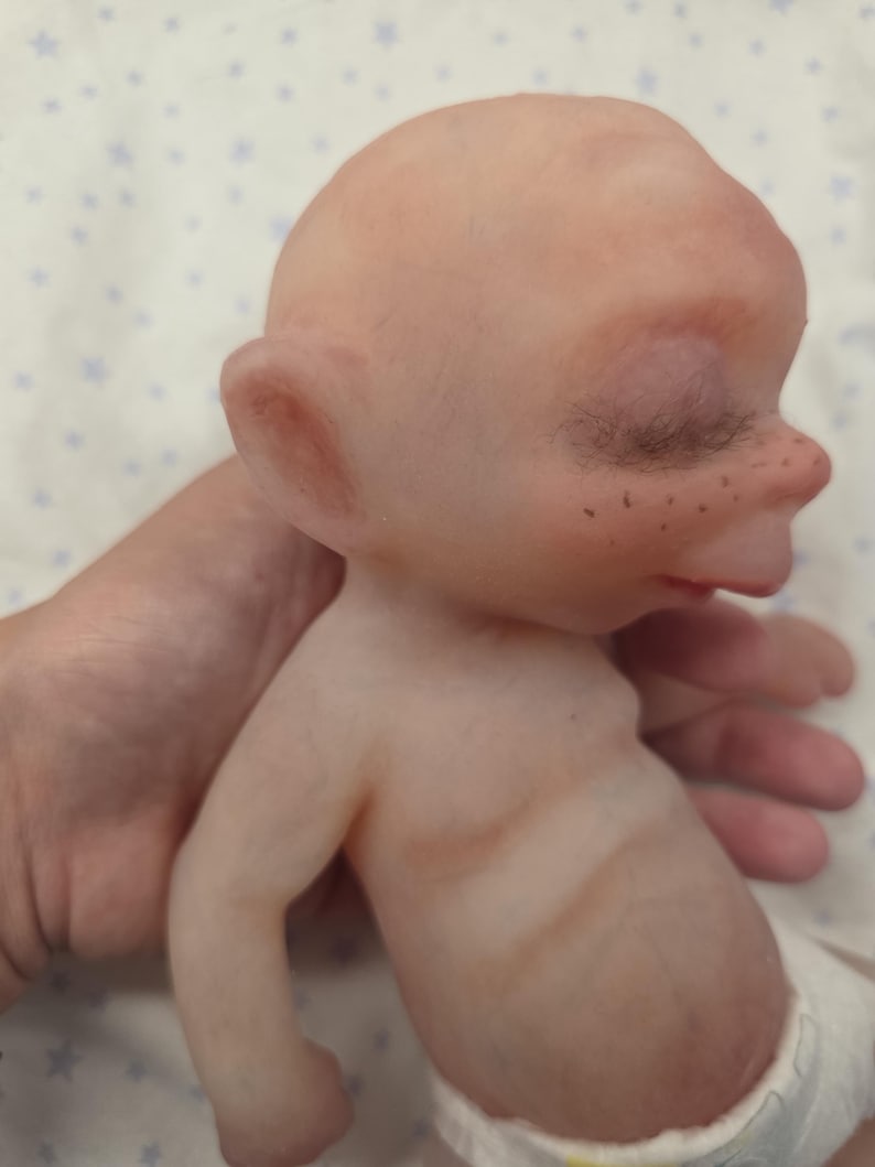 May include: A small, bald, pale-skinned doll with prominent ears, closed eyes with dark eyelashes, and visible freckles across the nose bridge. The doll has a rounded head, a small mouth, and a torso with subtle brown shading, wearing a white diaper.