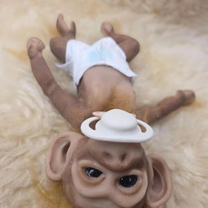 May include: A small monkey figurine with brown skin, dark eyes, and light brown fur around the neck. The monkey is wearing a white diaper and has a white pacifier in its mouth. The figurine is positioned in a lying down orientation.