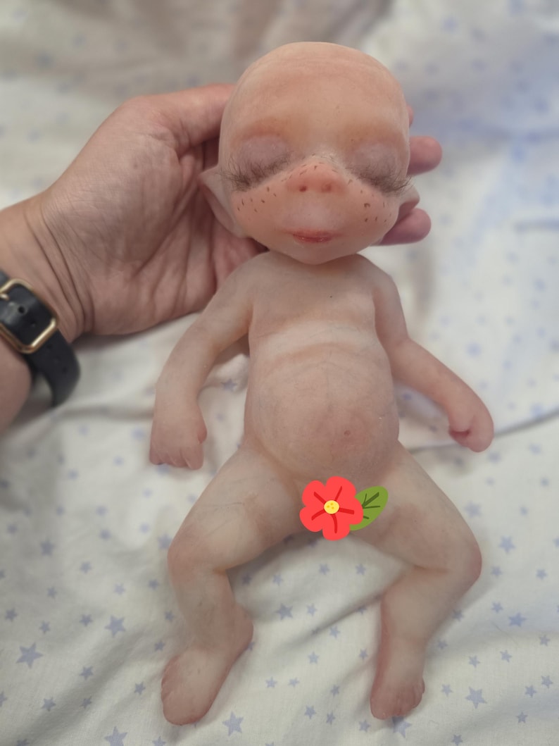May include: A small, pale, fantasy-style doll with a rounded head, pointed ears, and closed eyes featuring long, dark eyelashes. The doll has a smooth, light-toned body with subtle freckles across the nose and cheeks, and a red flower graphic.