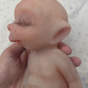 May include: A pale, smooth-skinned doll with a bald head, pointed ears, and closed eyes featuring long, dark eyelashes. The doll has a small nose, a defined mouth, and light brown freckles across the bridge of the nose and cheeks.