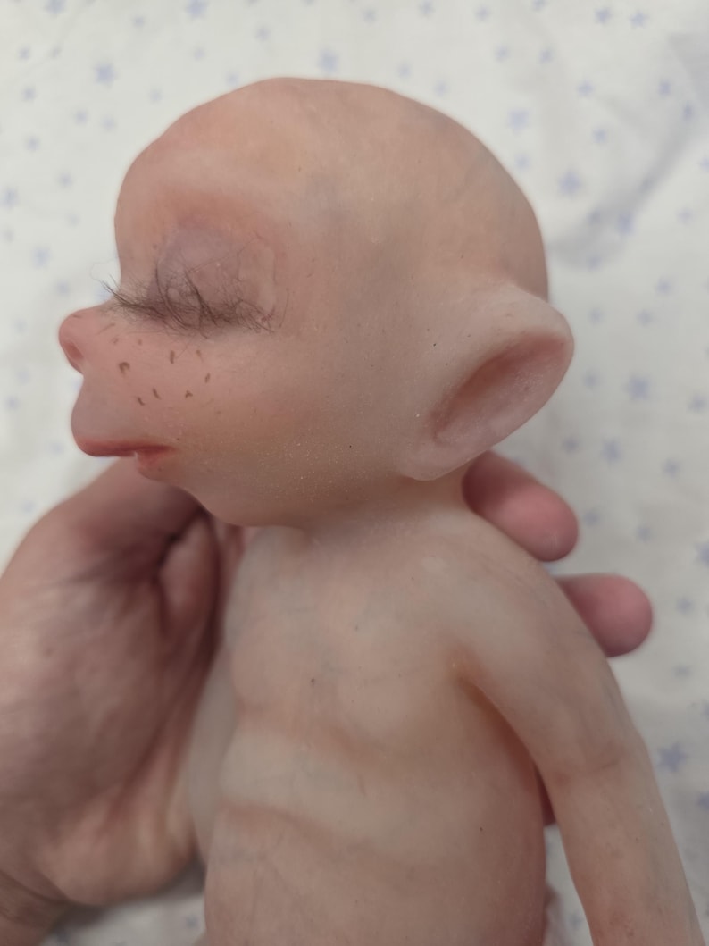 May include: A pale, smooth-skinned doll with a bald head, pointed ears, and closed eyes featuring long, dark eyelashes. The doll has a small nose, a defined mouth, and light brown freckles across the bridge of the nose and cheeks.
