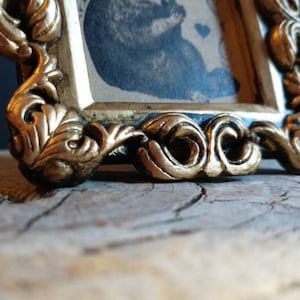 May include: An ornate picture frame with a gold-coloured finish and a black interior. The frame features a detailed, intricate design with a floral motif.