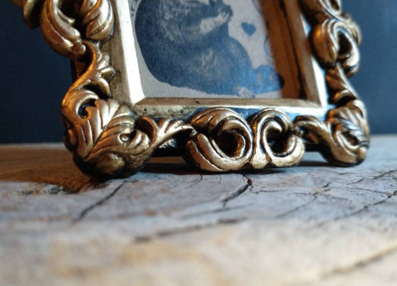 May include: An ornate picture frame with a gold-coloured finish and a black interior. The frame features a detailed, intricate design with a floral motif.