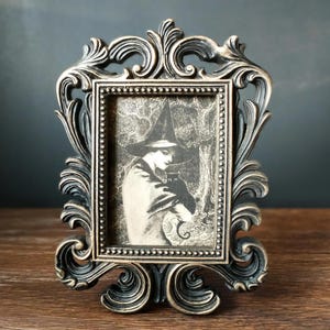 May include: A black and gold ornate picture frame with a black and white image of a witch holding a black cat.